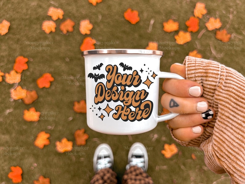 Puede incluir: Taza de camping de esmalte blanco con un dise&ntilde;o dorado y negro que dice "Your Design Here". La taza est&aacute; sostenida por una mano con un tatuaje en el dedo. El fondo es una imagen borrosa de hojas de oto&ntilde;o.