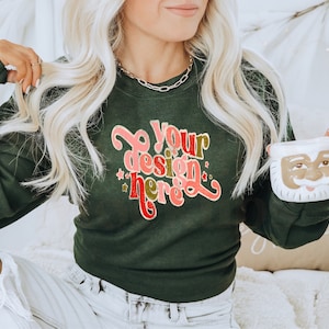 May include: A woman wearing a dark green sweatshirt with the text "Do your design here" in pink, red, and gold lettering. She is holding a white ceramic mug with a Santa Claus face.