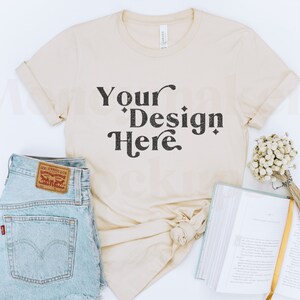 May include: A beige t-shirt with the text "Your Design Here" in black. The t-shirt is laid flat on a white surface with a pair of blue denim shorts, a book, and dried flowers.
