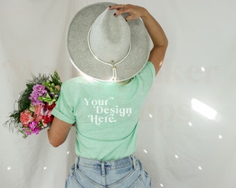 Puede incluir: Una persona lleva una camiseta verde claro con el texto "Your Design Here" en la espalda, un sombrero gris de ala ancha y unos vaqueros azules. La persona sostiene un ramo de flores rosas, rojas y blancas.