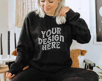 May include: Black crewneck sweatshirt with the text "YOUR DESIGN HERE" in a distressed white font. The sweatshirt is worn by a person sitting in a chair. The background includes decorative items and a wall hanging.