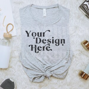 May include: A gray sleeveless shirt with the text "Your Design Here" printed on the front. The shirt is laid out on a white fluffy surface with a water bottle, two dumbbells, and a notebook.