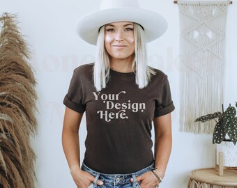 May include: A woman wearing a brown t-shirt with the text "Your Design Here" in a vintage font. She is wearing a white hat and blue jeans. The background is a white wall with a macrame wall hanging and a potted plant.