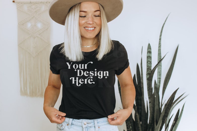 May include: A woman wearing a black t-shirt with the text "Your Design Here" in white. She is wearing a brown hat and has blonde hair.