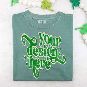 Może przedstawiać: Składana koszulka w kolorze turkusowym z napisem "Your design here" w stylu vintage. Koszulka leży na białym tle z biało-zieloną girlandą.