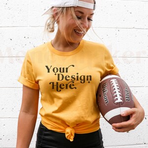 May include: Mustard yellow t-shirt with the text "Your Design Here" in black. The shirt is knotted at the waist. A brown and white Wilson football is held in the hands.