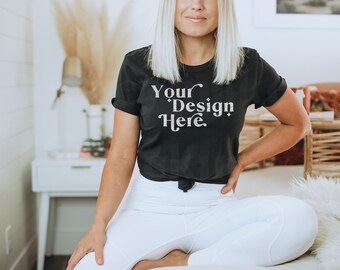 May include: A black t-shirt with the text "Your Design Here" in white lettering. The shirt is being worn by a person sitting on a white bed with a white fluffy throw.
