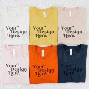 May include: Six folded t-shirts in different colours, including beige, yellow, peach, orange, and black. Each shirt has the text "Your Design Here" printed on the front.