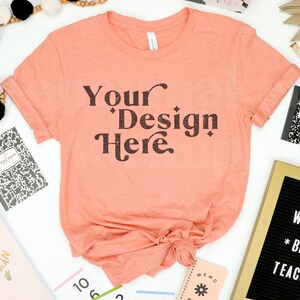 May include: A peach coloured t-shirt with the text "Your Design Here" in black. The shirt is laid flat on a white surface with school supplies around it, including a letter board that reads "Worlds Best Teacher".
