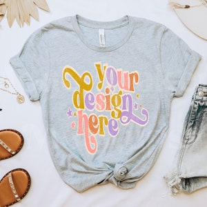May include: A gray t-shirt with the text "Your design here" in a retro font with colorful accents. The shirt is knotted at the bottom and is surrounded by accessories like sandals, sunglasses, a necklace, a hat, and denim shorts.