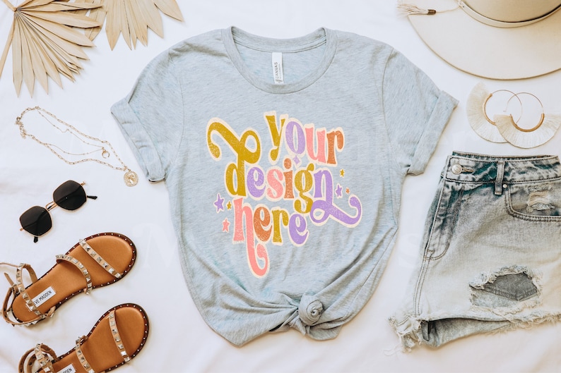 May include: A gray t-shirt with the text "Your design here" in a retro font with colorful accents. The shirt is knotted at the bottom and is surrounded by accessories like sandals, sunglasses, a necklace, a hat, and denim shorts.