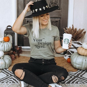 May include: A woman wearing a green t-shirt with the text "Your Design Here" sits on a rug with pumpkins and a cup of coffee. She is wearing a black hat with gold accents and black jeans.