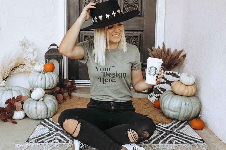 May include: A woman wearing a green t-shirt with the text "Your Design Here" sits on a rug with pumpkins and a cup of coffee. She is wearing a black hat with gold accents and black jeans.