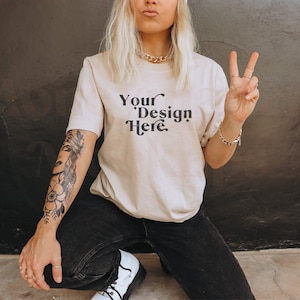 May include: A woman wearing a light beige t-shirt with the text "Your Design Here" printed on the front. She is wearing black jeans and has a tattoo on her arm.