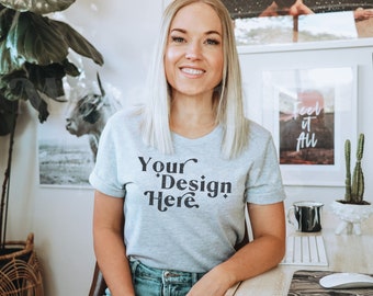 Peut inclure: Une femme portant un t-shirt gris avec le texte "Your Design Here" en noir. Elle est assise à un bureau avec un ordinateur, une tasse et un cactus.