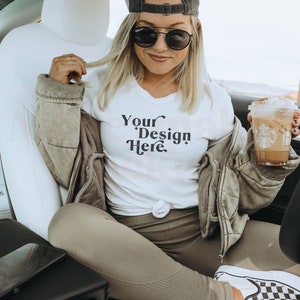 Pode incluir: Uma mulher vestindo uma camiseta branca com o texto "Your Design Here" em letras pretas, um boné de beisebol preto e calças marrons. Ela está segurando uma bebida de café em um copo de plástico transparente com tampa marrom.