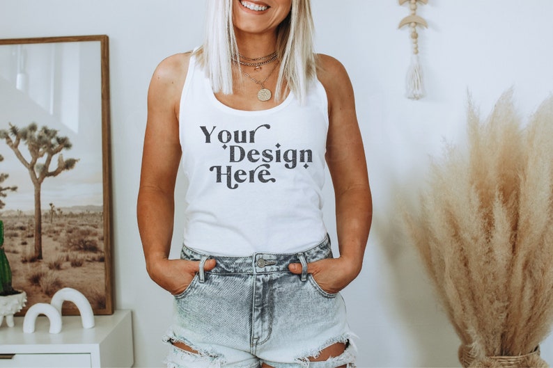 Puede incluir: Camiseta blanca de tirantes con el texto "Your Design Here" en negro. La camiseta est&aacute; siendo usada por una persona con el pelo largo y rubio. La persona lleva pantalones cortos de mezclilla azul claro.