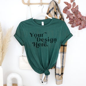May include: A dark green t-shirt with the text "Your Design Here" in black lettering. The t-shirt is hanging on a wooden rack with a plaid scarf and a white purse.