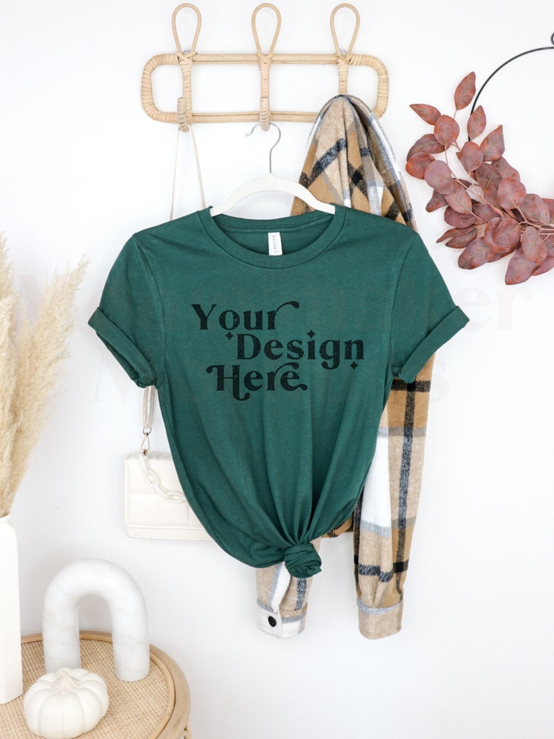 May include: A dark green t-shirt with the text "Your Design Here" in black lettering. The t-shirt is hanging on a wooden rack with a plaid scarf and a white purse.