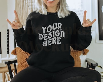 May include: A black sweatshirt with the text "YOUR DESIGN HERE" in white. The sweatshirt is being worn by a person with blonde hair, sitting on a chair with a brown pillow.