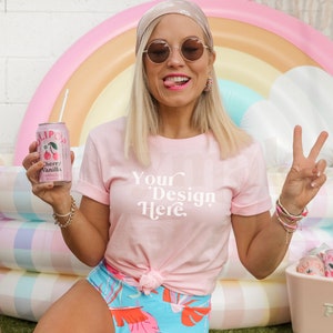 Könnte beinhalten: Eine Frau trägt ein pinkfarbenes T-Shirt mit dem Text "Your Design Here" in Weiß. Sie hält eine rosafarbene Dose Limonade mit einem Strohhalm und einer Kirsche auf dem Etikett. Sie sitzt vor einem regenbogenfarbenen aufblasbaren Pool-Schwimmreifen.