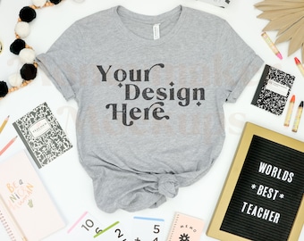 May include: A gray t-shirt with the text "Your Design Here" in black. The shirt is laid out on a white surface with school supplies around it, including a black letter board with the text "Worlds Best Teacher".
