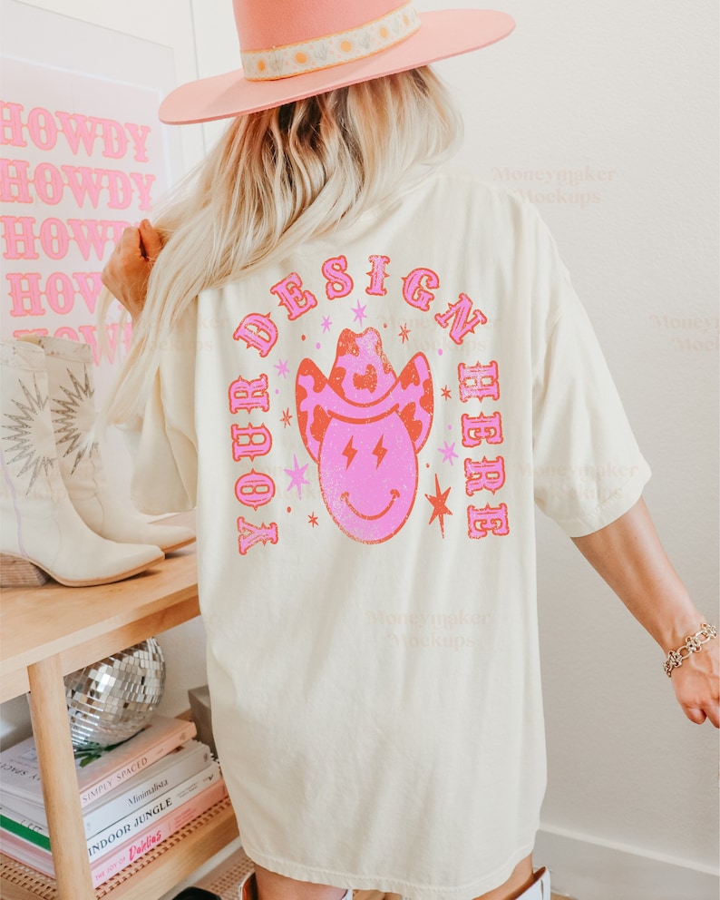 以下が含まれることがあります： 薄いベージュのTシャツで、背面にピンクとオレンジのデザインが施されています。デザインは、笑顔と稲妻が描かれた漫画の牛仔帽です。「YOUR DESIGN HERE」というテキストが、牛仔帽の周りを円形に囲んでいます。