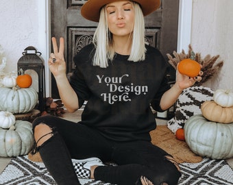 May include: Black sweatshirt with the text "Your Design Here". The person is wearing a brown hat and black ripped jeans. There are pumpkins and decorative gourds in the background, creating a fall theme.