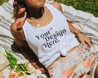 May include: A woman wearing a white tank top with the text "Your Design Here" printed on it, a floral patterned skirt, and a pink bandana. She is lying on a striped blanket in a grassy area.