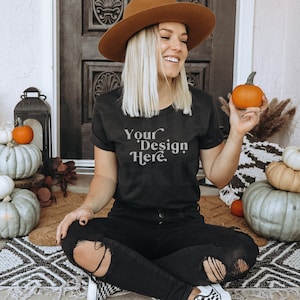 May include: A woman wearing a black t-shirt with the text "Your Design Here" sitting on a patterned rug with pumpkins around her. She is holding a small orange pumpkin in her hand.