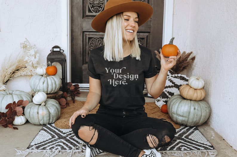 May include: A woman wearing a black t-shirt with the text "Your Design Here" sitting on a patterned rug with pumpkins around her. She is holding a small orange pumpkin in her hand.