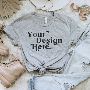 Puede incluir: Una camiseta gris con el texto "Your Design Here" impreso en la parte delantera. La camiseta está anudada en la parte delantera y se combina con unos vaqueros azul claro y unas botas marrones.