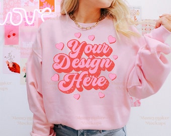 Puede incluir: Una sudadera rosa con un gráfico de estilo retro que dice "Your Design Here" en letras rojas y rosas con corazones. La sudadera está siendo modelada por una persona que lleva jeans azules.