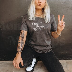 Może przedstawiać: Kobieta w ciemnoszarym t-shircie z nadrukiem "Your Design Here". Klęczy przed ciemnoszarą ścianą i ma tatuaż na ramieniu.