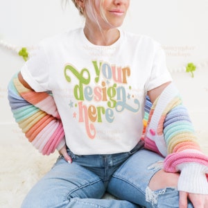 Puede incluir: Una camiseta blanca con un diseño retro que dice "Your design here" en letras de colores. La camiseta la lleva puesta una persona sentada con una chaqueta de punto arcoíris.