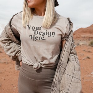 May include: A woman wearing a light brown t-shirt with the text "Your Design Here" in black. She is standing in a desert landscape with red rock formations in the background.