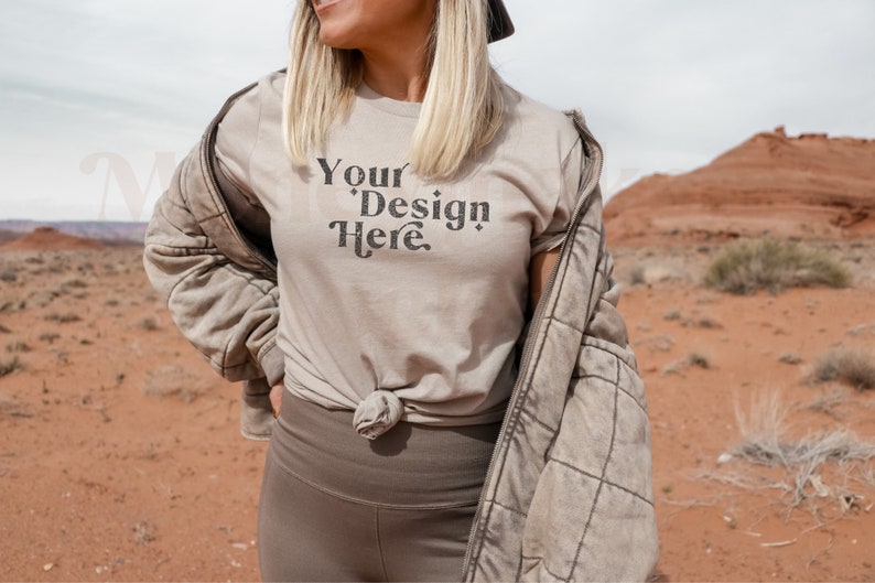 May include: A woman wearing a light brown t-shirt with the text "Your Design Here" in black. She is standing in a desert landscape with red rock formations in the background.