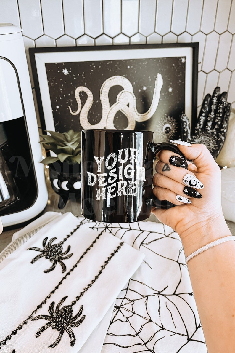K&ouml;nnte beinhalten: Schwarze Kaffeetasse mit dem Text "YOUR DESIGN HERE" in wei&szlig;en Buchstaben. Die Tasse wird von einer Hand mit schwarz-wei&szlig;em Nagellack gehalten.