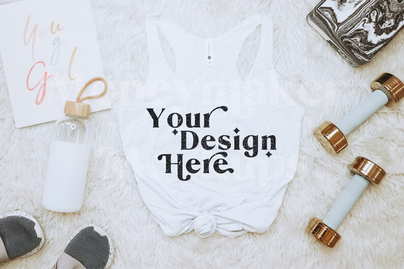 Może przedstawiać: Biały top bez rękaw&oacute;w z napisem "Your Design Here" czarnym pismem jest centralnym punktem tego zdjęcia. Obraz zawiera r&oacute;wnież butelkę wody, hantle, notatnik i trampki, wszystko ułożone na białej, puszystej powierzchni.