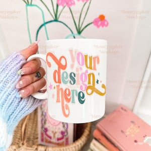 Op de afbeelding: Een witte keramische mok met een kleurrijk ontwerp met de tekst "Your design here" in een retro-lettertype. De mok wordt vastgehouden door een hand met een blauw-witte gebreide handschoen.