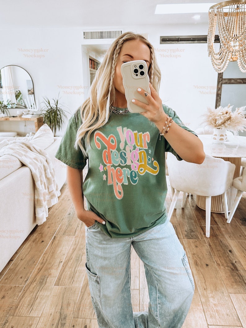 May include: An olive green t-shirt with the text "Your design here" in a colorful, retro font. The shirt is paired with light wash denim cargo pants. The person is holding a phone with a light-colored case.