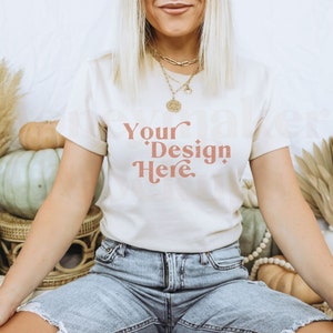 May include: A woman wearing a white t-shirt with the text "Your Design Here" in a peachy-pink colour. She is sitting on a white surface with pumpkins and other autumnal decor around her.