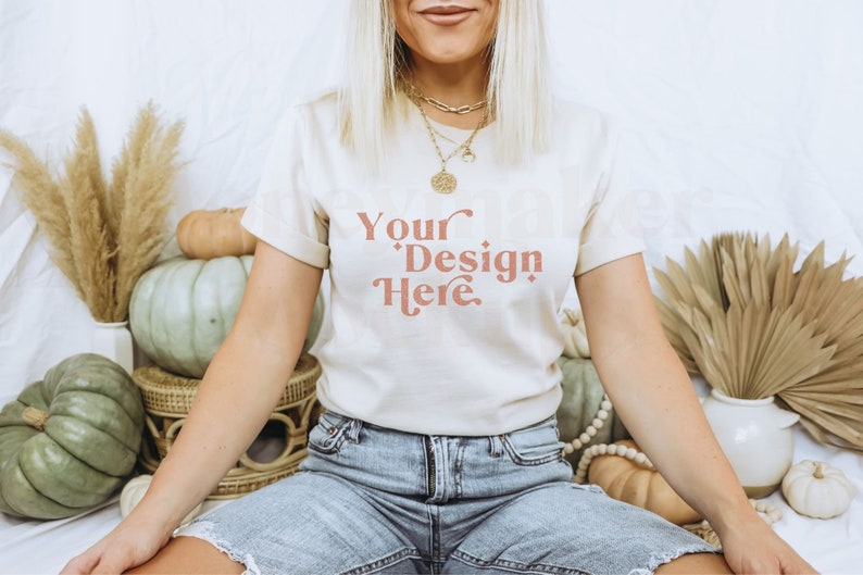 May include: A woman wearing a white t-shirt with the text "Your Design Here" in a peachy-pink colour. She is sitting on a white surface with pumpkins and other autumnal decor around her.