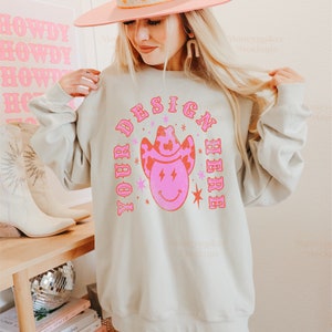 May include: A light grey sweatshirt with a pink and red design that says "YOUR DESIGN HERE" with a cartoon cowboy hat and smiley face. The model is wearing a pink cowboy hat and white cowboy boots.