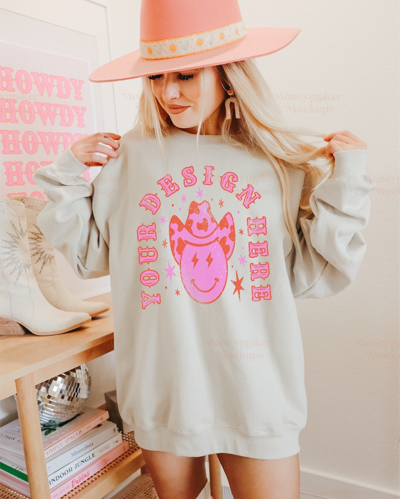May include: A light grey sweatshirt with a pink and red design that says "YOUR DESIGN HERE" with a cartoon cowboy hat and smiley face. The model is wearing a pink cowboy hat and white cowboy boots.