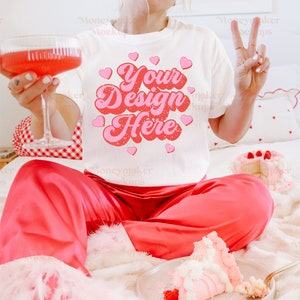 Op de afbeelding: Een wit T-shirt met een roze en rode afbeelding waarop "Your Design Here" staat, omringd door roze hartjes. Het shirt wordt gedragen door iemand die op een wit, pluche bed zit met een stuk taart op een bord.