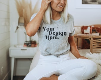 Peut inclure: Une femme portant un t-shirt gris avec le texte "Your Design Here" en lettres noires. Elle est assise sur un lit avec un pantalon blanc et un jeté en fourrure blanche.
