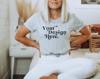 Peut inclure: Une femme portant un t-shirt gris avec le texte "Your Design Here" en noir. Elle est assise sur un lit blanc, les jambes croisées.