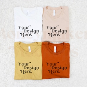 May include: Four folded t-shirts in different colours: white, beige, mustard yellow, and orange. Each shirt has the text "Your Design Here" printed on the front.