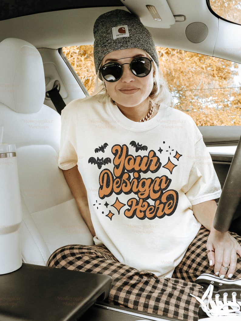 May include: A white t-shirt with a retro design featuring bats, stars, and the text "Your Design Here". The shirt is being worn by a person in a car.